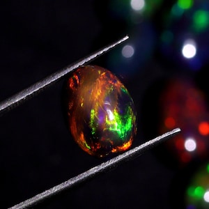 Pode incluir: Close-up de uma gema de opala escura e iridescente com flashes de verde, laranja e roxo. A opala &eacute; segurada por uma pin&ccedil;a contra um fundo desfocado de outras opalas.