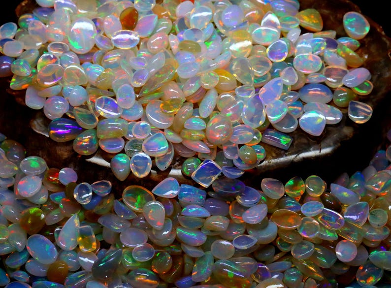 Op de afbeelding: Een verzameling opalen in verschillende vormen en maten, met een scala aan kleuren, waaronder wit, blauw, groen en oranje. De edelstenen hebben een parelmoerglans, die het licht reflecteert en een glinsterend effect cre&euml;ert. De opalen zijn verspreid.