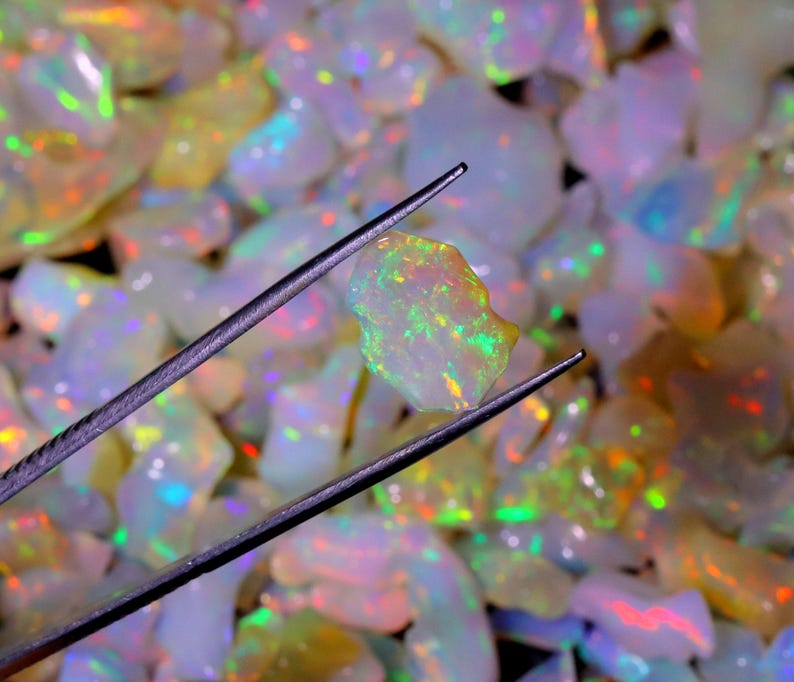 May include: A close-up of a white opal gemstone with iridescent green, yellow, and orange flashes. The opal is held by tweezers and surrounded by a pile of other opals.