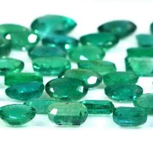 May include: A collection of emerald green gemstones, cut and polished into oval shapes. The stones are arranged in a row on a white surface.