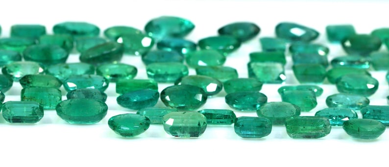 May include: A collection of emerald green gemstones, cut and polished into oval shapes. The stones are arranged in a row on a white surface.