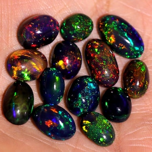 以下が含まれることがあります： 緑、青、赤、金色の虹色の光沢を持つ、13個の楕円形のブラックオパルのコレクション。オパールは手のひらに並べられています。