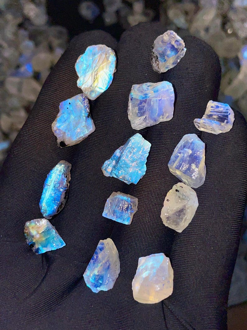 May include: A collection of raw moonstone crystals, displaying a range of blue, white, and iridescent colors. The gemstones are irregularly shaped and vary in size, with a shimmering, translucent quality. The crystals are held against a dark background.