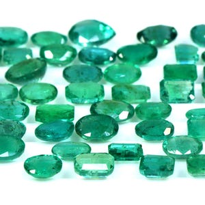 May include: A collection of emerald green gemstones, cut into various shapes and sizes, including oval, rectangular, and square. The gemstones are arranged in a row on a white background.