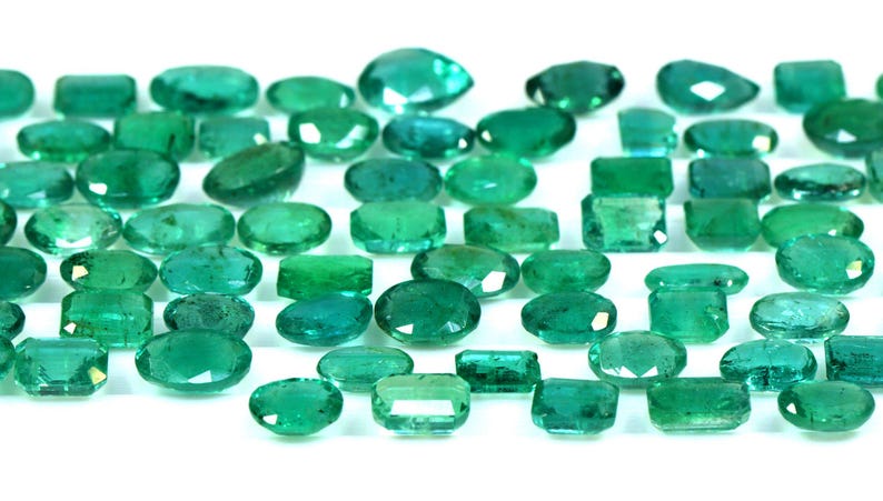May include: A collection of emerald green gemstones, cut into various shapes and sizes, including oval, rectangular, and square. The gemstones are arranged in a row on a white background.