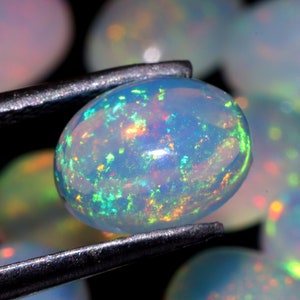 Könnte beinhalten: Nahaufnahme eines weißen Opals mit schillernden grünen, blauen und orangen Farbtönen. Der Opal ist oval geformt und wird mit einer Pinzette gehalten.