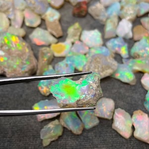 Op de afbeelding: Een verzameling ruwe opaal edelstenen, sommige met iriserende kleuren, worden getoond op een grijs oppervlak. Een opaal, met een groene en blauwe iriserende glans, wordt vastgehouden met een pincet.