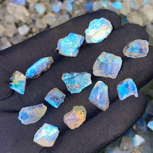 May include: A collection of raw, iridescent moonstone crystals. The stones display a range of blues, whites, and hints of yellow and orange. The crystals are held against a black background, highlighting their natural beauty and unique shapes.