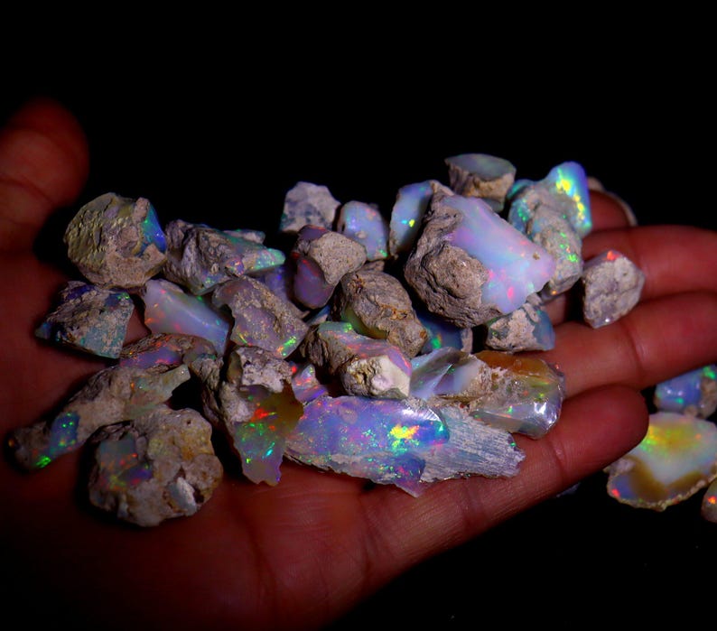 May include: A collection of rough opal stones, displaying a spectrum of colours such as blues, greens, and iridescent flashes. The opals are held against a dark backdrop, accentuating their natural texture and beauty.
