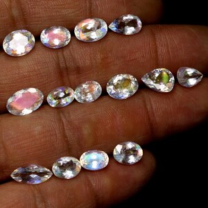 May include: A collection of iridescent gemstones in various shapes, including oval and pear cuts. The gems display a range of colors, with flashes of pink, blue, and yellow. The stones are held in a hand against a dark background, showcasing their brilliance.