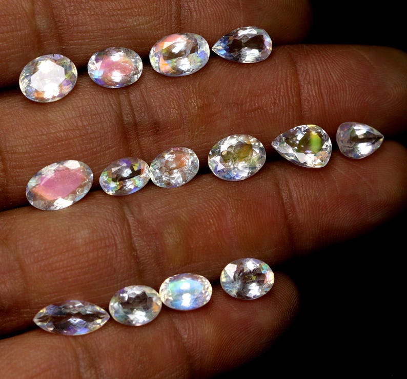 May include: A collection of iridescent gemstones in various shapes, including oval and pear cuts. The gems display a range of colors, with flashes of pink, blue, and yellow. The stones are held in a hand against a dark background, showcasing their brilliance.