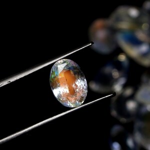 May include: An oval-cut gemstone held by tweezers against a black background. The gem has a mix of colors, including orange, yellow, and blue, with a clear, reflective surface. The gem is the focal point, with other gems blurred in the background.