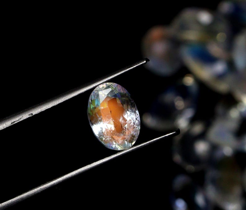 May include: An oval-cut gemstone held by tweezers against a black background. The gem has a mix of colors, including orange, yellow, and blue, with a clear, reflective surface. The gem is the focal point, with other gems blurred in the background.