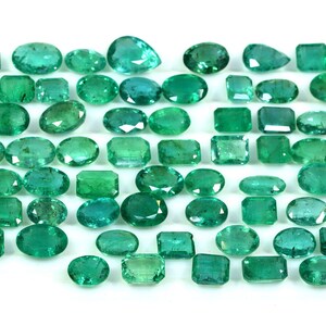May include: A collection of green emerald gemstones of various shapes and sizes. The emeralds are arranged on a white surface.