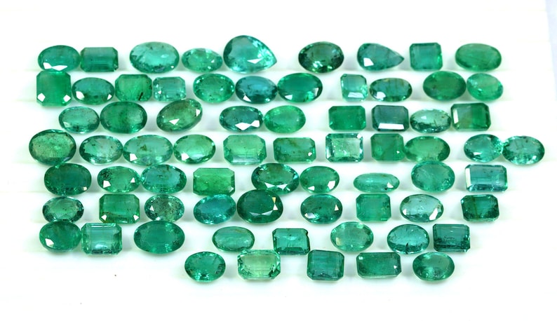 May include: A collection of green emerald gemstones of various shapes and sizes. The emeralds are arranged on a white surface.