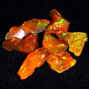 May include: A group of seven orange and green opal gemstones. The opals are rough and have an iridescent sheen.