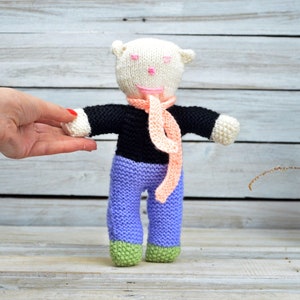 May include: A white knitted toy bear wearing a black sweater, pink scarf, and purple pants. The bear has a pink nose and pink eyes.