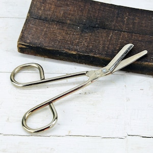 May include: Silver-toned metal scissors with long handles and a pointed tip. The scissors are lying on a white wooden surface.
