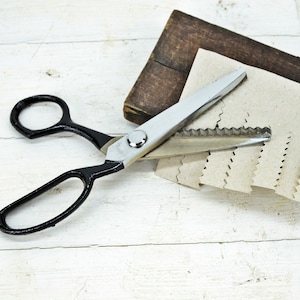 May include: A pair of black-handled pinking shears with a silver blade on a white wooden surface. The shears are cutting a stack of brown paper.