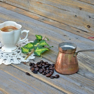 May include: A white coffee cup with a lace doily, a copper coffee pot, and a pile of coffee beans on a wooden surface.