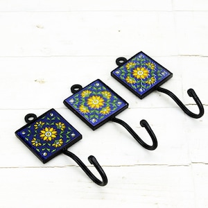 Könnte beinhalten: Drei dekorative Wandhaken mit blauen und gelben floralen Fliesenmustern. Jeder Haken hat eine schwarze Metallbasis und einen einzelnen Haken.