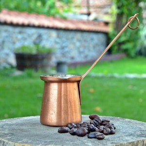 Könnte beinhalten: Eine türkische Kaffeekanne aus Kupfer mit langem Henkel steht auf einer Holzoberfläche neben einem Haufen Kaffeebohnen.