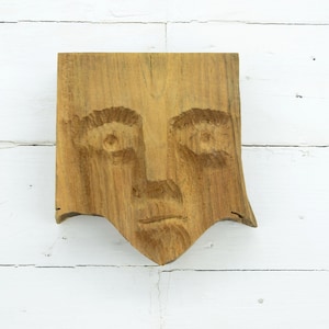 May include: A wooden sculpture of a face with a carved expression. The face is carved from a single piece of wood and has a natural wood finish.