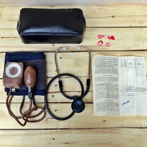 Puede incluir: Un bolso médico de cuero negro, un estetoscopio, un brazalete de presión arterial, un esfigmomanómetro y un folleto de instrucciones médicas sobre una superficie de madera.
