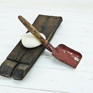 Peut inclure: Nature morte rustique avec une petite pelle rouge usée avec un manche en bois, posée sur une poutre en bois sombre et vieillie. Une pierre blanche et lisse est également présente.
