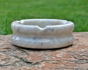 May include: A round, light gray marble ashtray with a double-banded design. The ashtray sits on a rough, brown stone surface, with a blurred green background. The ashtray has a single notch for resting a cigarette.