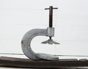 May include: A silver-coloured metal C-clamp with a threaded rod and handle, set on a dark brown, weathered wooden base. The background is a white wooden surface. The clamp is a tool used for woodworking.
