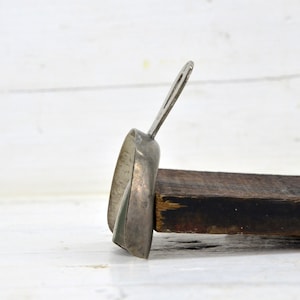 Può includere: Una paletta in metallo argentato con un manico in legno. La paletta ha la forma di un semicerchio e ha una piccola maniglia sul lato. La paletta è appoggiata su un pezzo di legno.