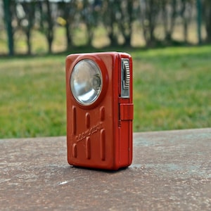 Puede incluir: Una linterna de metal roja con una lente plateada y una perilla plateada en el lateral. La linterna está sobre una superficie de metal oxidado.