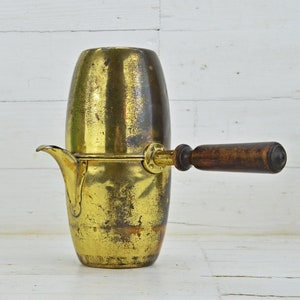 Puede incluir: Una cafetera de latón vintage con un mango de madera. La cafetera tiene un aspecto desgastado y antiguo y está hecha de dos piezas que encajan.