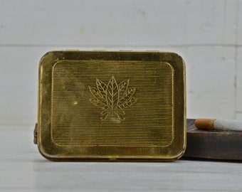 Puede incluir: Un estuche de metal dorado para cigarrillos con un diseño de hoja en la parte delantera.