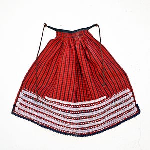 Peut inclure: Un tablier à carreaux rouges et noirs doté d'une taille froncée avec de fines attaches sombres. La partie inférieure est ornée de quatre rangées horizontales de dentelle blanche au crochet, chacune agrémentée de petites perles rouges et blanches.