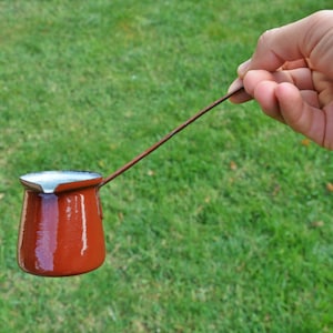 Puede incluir: Una cafetera de metal roja con un mango largo, llena de líquido blanco, sostenida por una mano sobre un fondo de hierba verde.