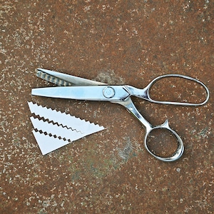 Puede incluir: Tijeras decorativas plateadas con un diseño de hoja único, junto a un trozo de papel blanco cortado. Las tijeras tienen un asa de bucle y un orificio para el dedo más pequeño. El fondo es una superficie texturizada de color marrón oxidado.