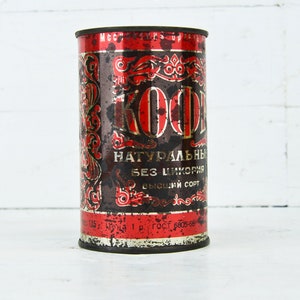 May include: A vintage red and black metal can with the word "KOФE" in gold lettering. The can is labeled "НАТУРАЛЬНЫЙ БЕЗ ЦИКОРИЯ ВЫСШИЙ СОРТ" in Cyrillic script.