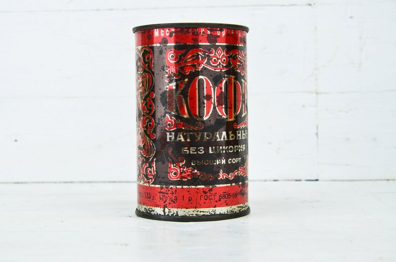 May include: A vintage red and black metal can with the word "KOФE" in gold lettering. The can is labeled "НАТУРАЛЬНЫЙ БЕЗ ЦИКОРИЯ ВЫСШИЙ СОРТ" in Cyrillic script.