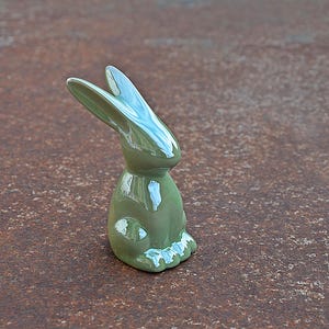 Op de afbeelding: Een glanzend, saliegroen keramiek konijnbeeldje. Het konijn zit met zijn oren omhoog. Het beeldje heeft een glad, reflecterend oppervlak en is een decoratief item.