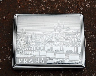 May include: Silver rectangular cigarette case with an engraved cityscape of Prague. The case features a detailed depiction of buildings, bridges, and the word "PRAHA" at the bottom. The case is on a textured, brown surface.