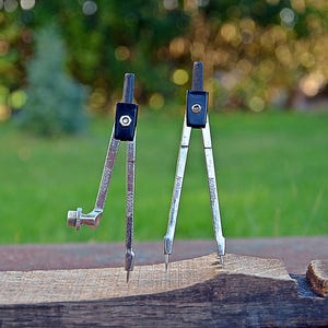 May include: Two silver compasses with black accents are positioned on a wooden surface. The compasses have sharp points and are set against a blurred green background. The tools are used for drawing circles and arcs.