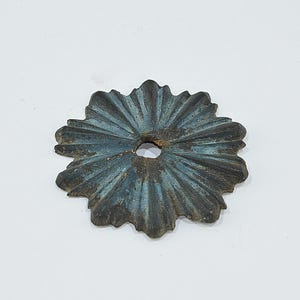 Op de afbeelding: Een verweerd, bloemvormig metalen decoratief stuk met een centraal gat. Het stuk heeft een donkere, verouderde uitstraling met blauwgroene en bruine tinten. Het ontwerp heeft stralende lijnen, die een bloemen- of zonnestraalmotief suggereren.