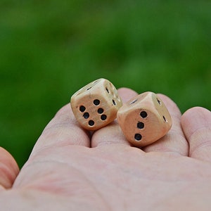 Puede incluir: Dos dados de madera con puntos negros en una mano. Los dados son de color marrón claro con veta de madera natural.