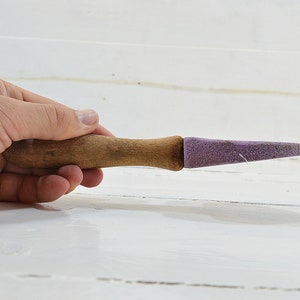 Puede incluir: Una herramienta de afilado con mango de madera y una piedra de afilar de color morado. La herramienta se sostiene en una mano.