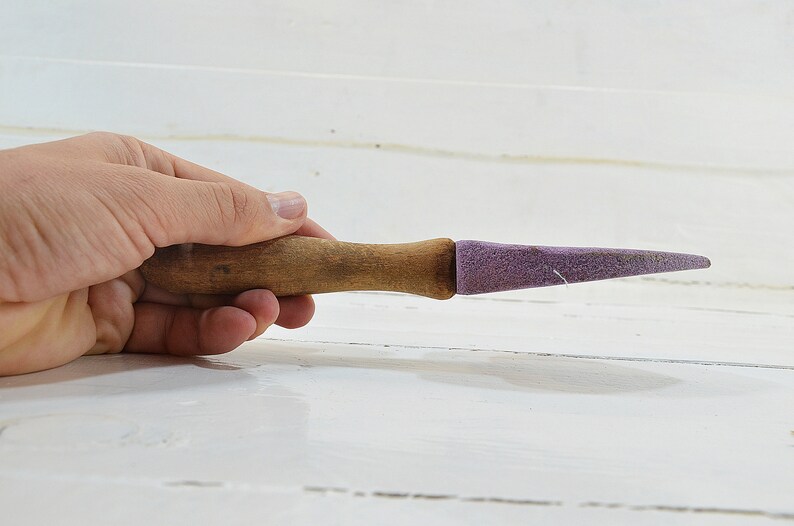 Puede incluir: Una herramienta de afilado con mango de madera y una piedra de afilar de color morado. La herramienta se sostiene en una mano.