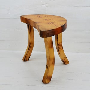 May include: A wooden stool with a half-moon shaped seat and three curved legs. The stool is made of natural wood with a light brown finish.