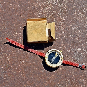 Könnte beinhalten: Ein Vintage-Kompass mit rotem Lederarmband und einer kleinen, offenen Pappschachtel. Der Kompass hat ein goldfarbenes Gehäuse, ein schwarzes Zifferblatt und eine silberne Nadel. Das Armband ist etwa 1,3 cm breit.