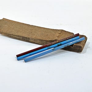Può includere: Tre matite in tonalità di blu e una matita rosso scuro poggiano su un pezzo di legno stagionato. Le matite sono disposte diagonalmente, con il pezzo di legno che funge da base rustica su uno sfondo bianco.